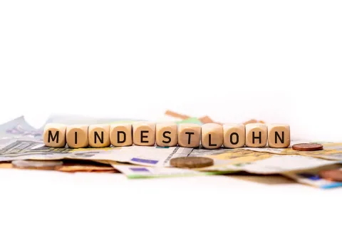 Holzwürfel bilden das Wort Mindestlohn auf einfarbigem, weißem Hintergrund mit Banknoten und Münzen