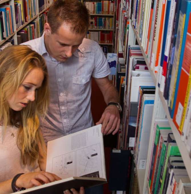 Zwei Studenten stehen zwischen Bücherregalen in einer Bibliothek und lesen in einem Buch. 
