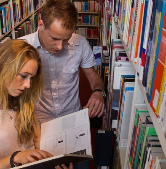 Zwei Studenten stehen zwischen Bücherregalen in einer Bibliothek und lesen in einem Buch.