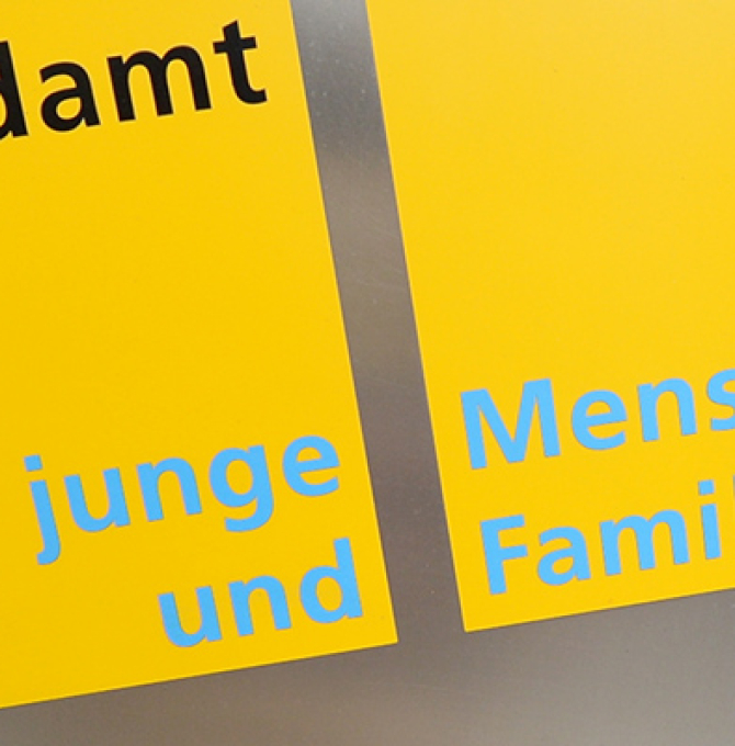 Das Bild zeigt ein gelbes Schild an einer Hauswand auf dem steht: "Jugendamt. Für junge Menschen und Familien" Rechts daneben befindet sich eine Straße mit parkenden Autos. 