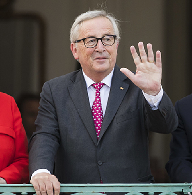 Das Bild zeigt von links: Bundeskanzlerin Angela Merkel in einem roten Sakko, EU-Kommissionspräsident Jean-Claude Juncker in einem dunkelgrauen Anzug mit violetter Krawatte und ganz rechts frankreichs Präsident Emmanuel Macron in einem dunklen Anzug mit schwarzer Krawatte. Alle drei stehen hinter einer grünen Brüstung auf Schloss Meseberg und schauen Richtung Kameras. Juncker winkt. 